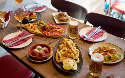 A wooden table infront of a window with sun shining through. Lots of small plates of Spanish food and glasses of beer are on the table. Including a large platter of calamari, antipasti board and dish of meatballs.