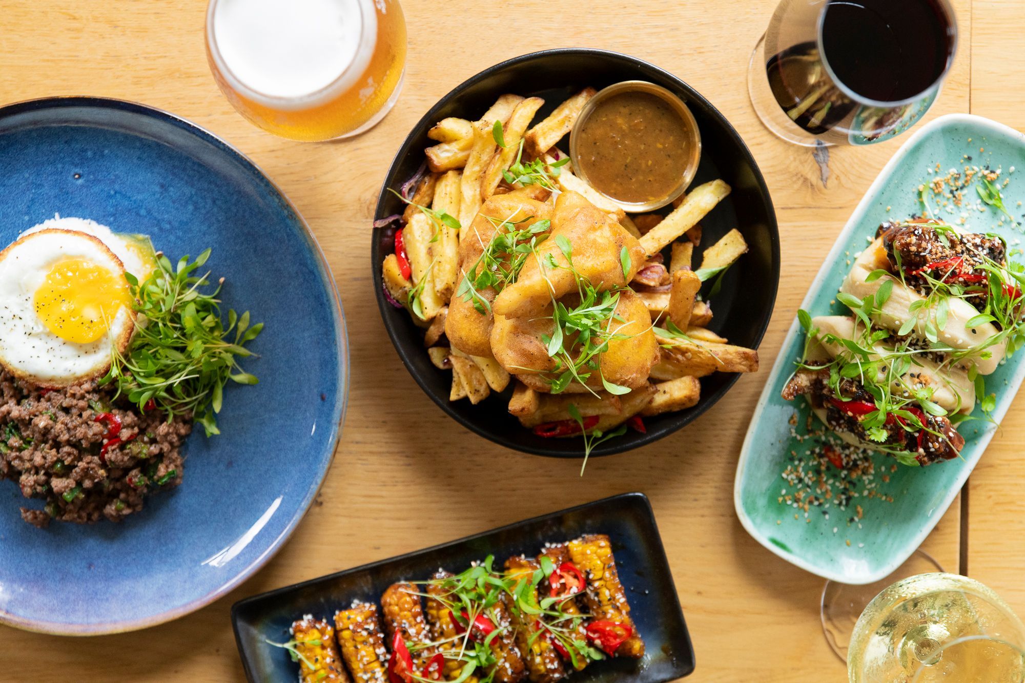 over head shot of the dishes of The Stirling Arms menu, four different dishes including meat with egg, bao buns, fries with sauce, and cooked corn with spices.