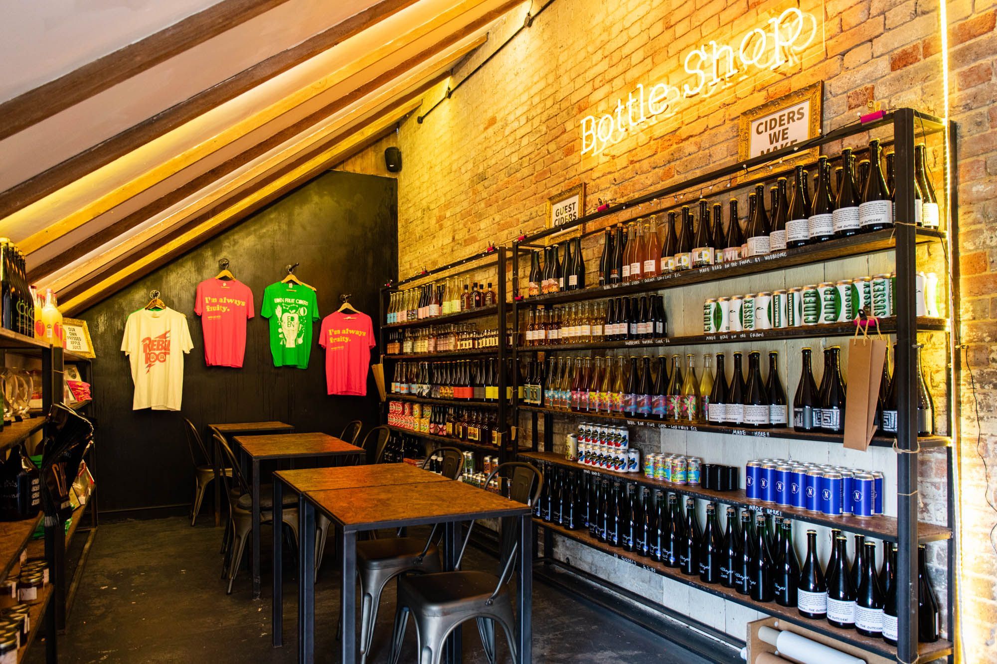 The Cider Tap Sussex Bottle Shop part of the restaurant, black shelf suit filled with various alcoholic drinks, white, pink and green shirts hanging on the wall