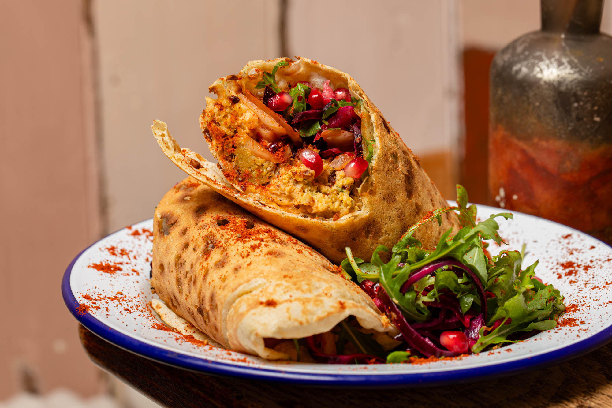 Close-up of a vibrant, freshly made wrap cut in half to reveal a colourful filling of spiced chickpea, beetroot slaw, rocket, onions, and pomegranate seeds, all dusted with paprika. Served on a white enamel plate with a fresh salad garnish.