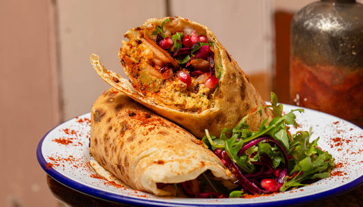 Close-up of a vibrant, freshly made wrap cut in half to reveal a colourful filling of spiced chickpea, beetroot slaw, rocket, onions, and pomegranate seeds, all dusted with paprika. Served on a white enamel plate with a fresh salad garnish.