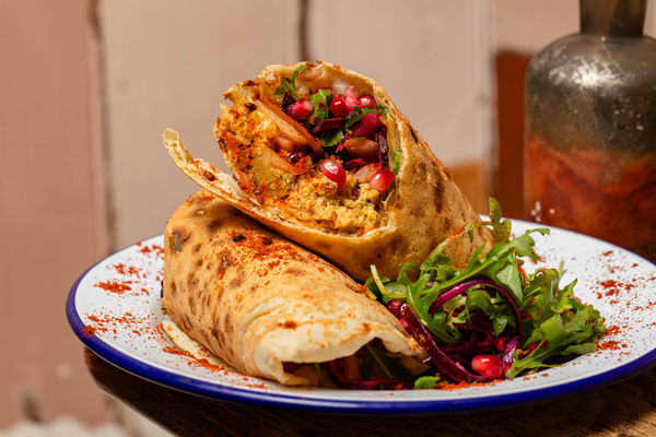 Close-up of a vibrant, freshly made wrap cut in half to reveal a colourful filling of spiced chickpea, beetroot slaw, rocket, onions, and pomegranate seeds, all dusted with paprika. Served on a white enamel plate with a fresh salad garnish.