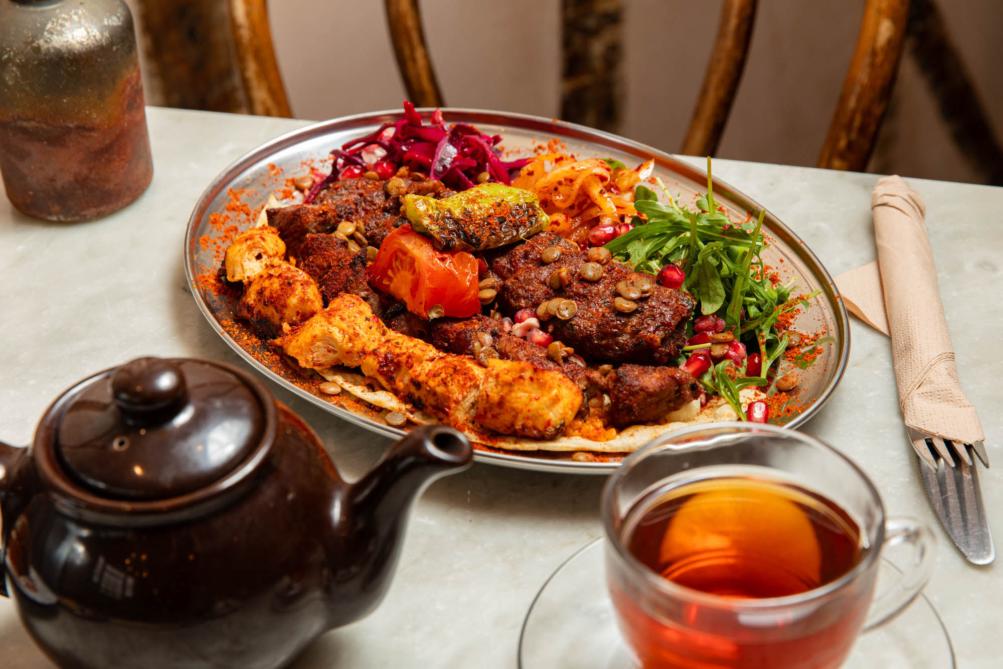A vibrant Middle Eastern mixed grill platter featuring spiced chicken skewers, grilled kofta, roasted vegetables, lentils, fresh rocket, pickled slaw, and pomegranate seeds, served on flatbread. Accompanied by a glass of tea and a dark teapot, set on a marble table with rustic wooden chairs in the background.