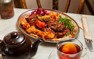 A vibrant Middle Eastern mixed grill platter featuring spiced chicken skewers, grilled kofta, roasted vegetables, lentils, fresh rocket, pickled slaw, and pomegranate seeds, served on flatbread. Accompanied by a glass of tea and a dark teapot, set on a marble table with rustic wooden chairs in the background.