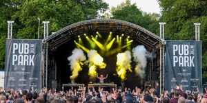 Picture features pub in the park at Preston Park. A man is jumping on stage in front of thousands of fans as pub in the park begins. There are stage lights and dry ice in the background.