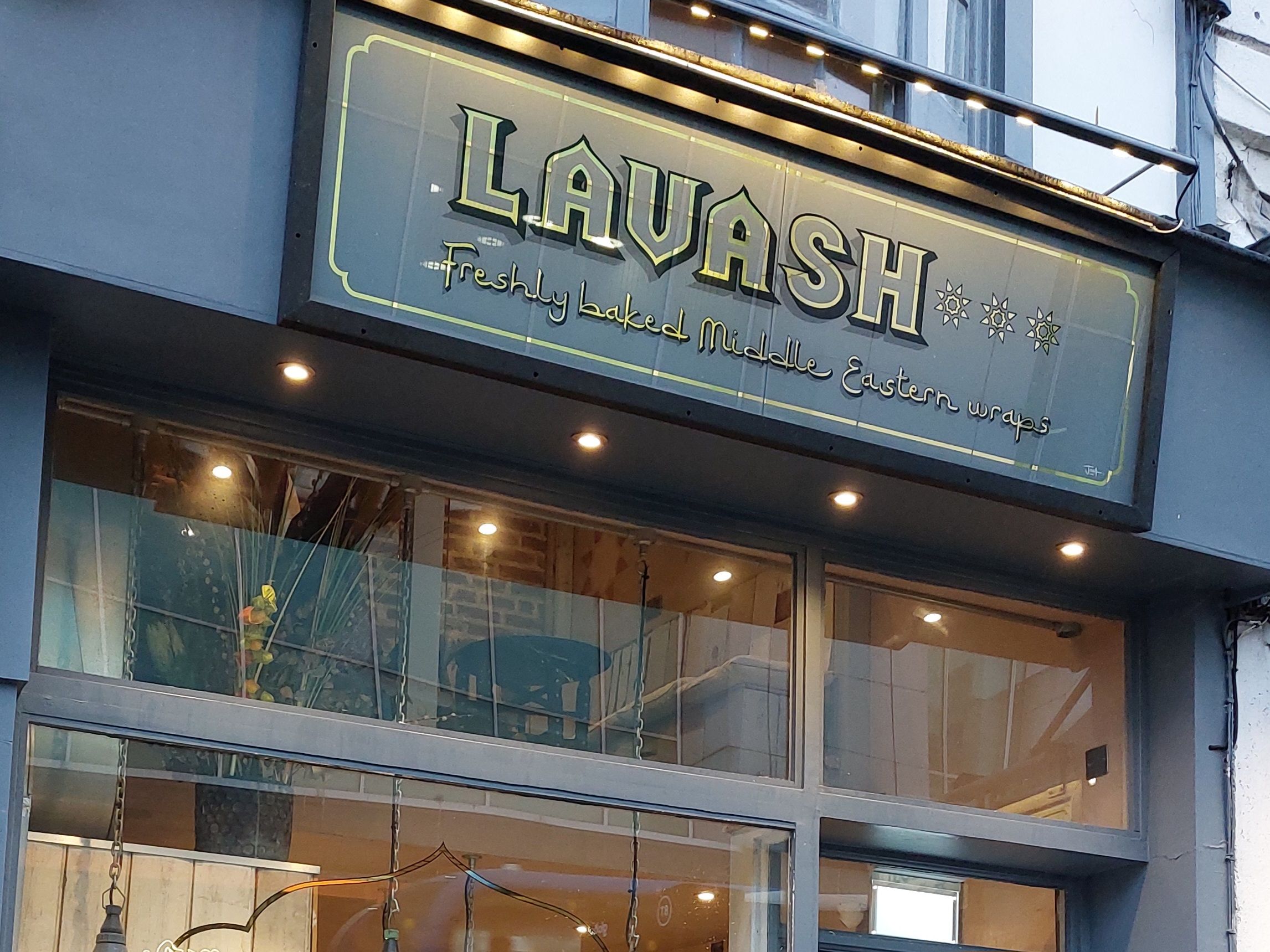 A sign over a restaurant window is written in an Arabic style font and reads LAVASH Freshly baked Middle Eastern Wraps
