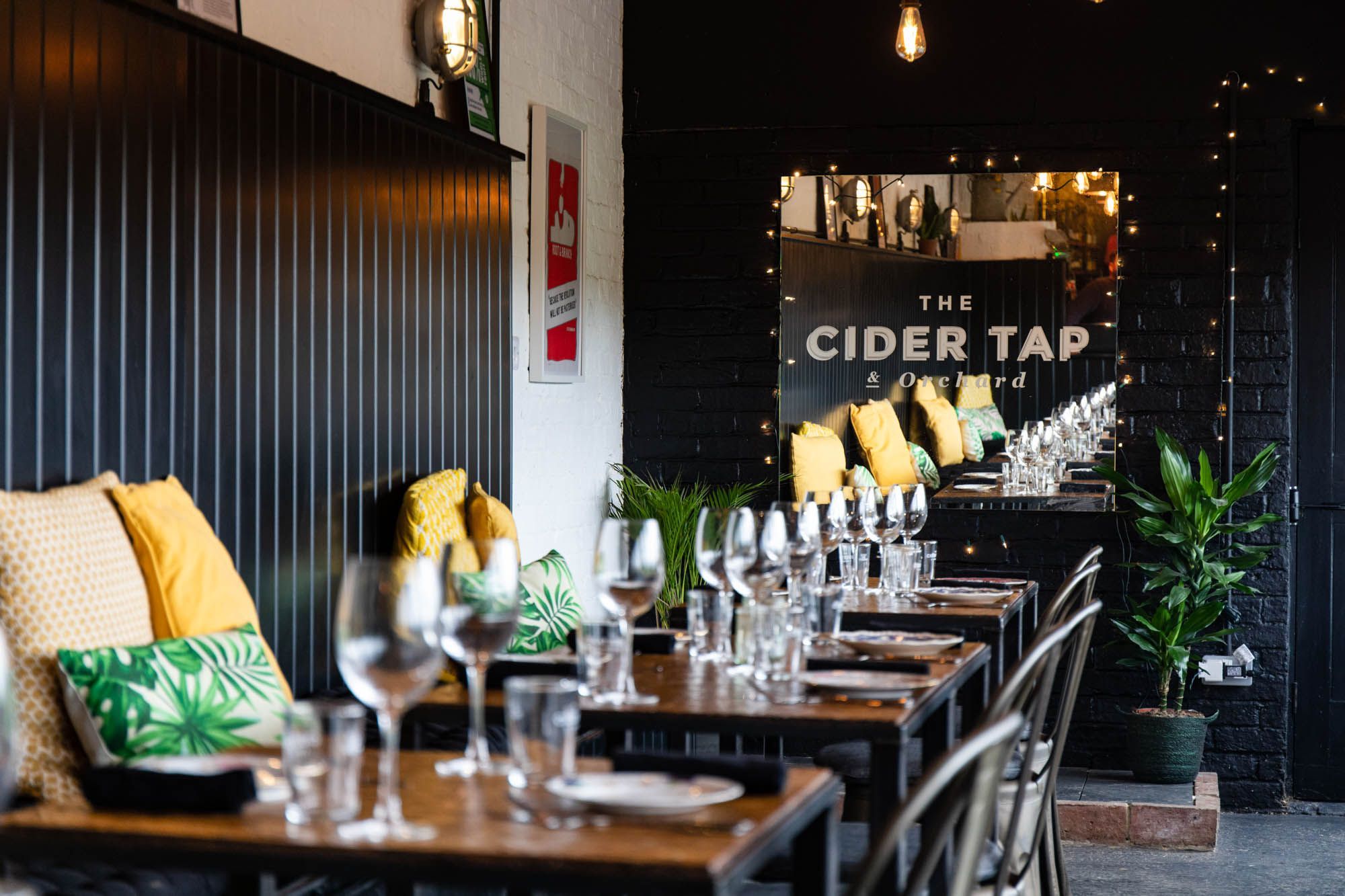 Interior of The Cider Tap, black and white walls, brown tables laid out with glasses for drinks, black chairs and yellow pillows