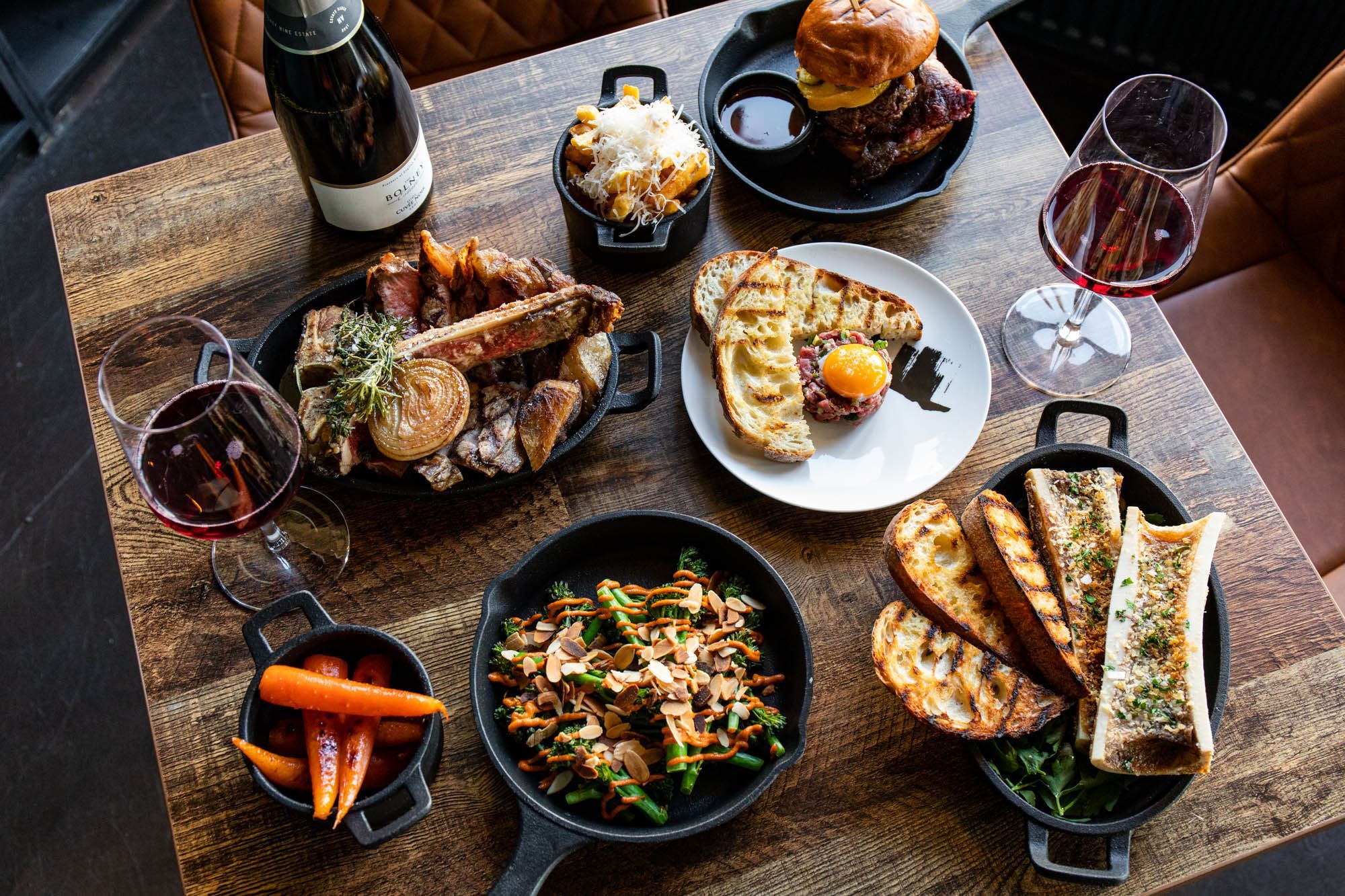 over head shot of the brown table laid out with couple of different main meat dishes and sides, served with red wine