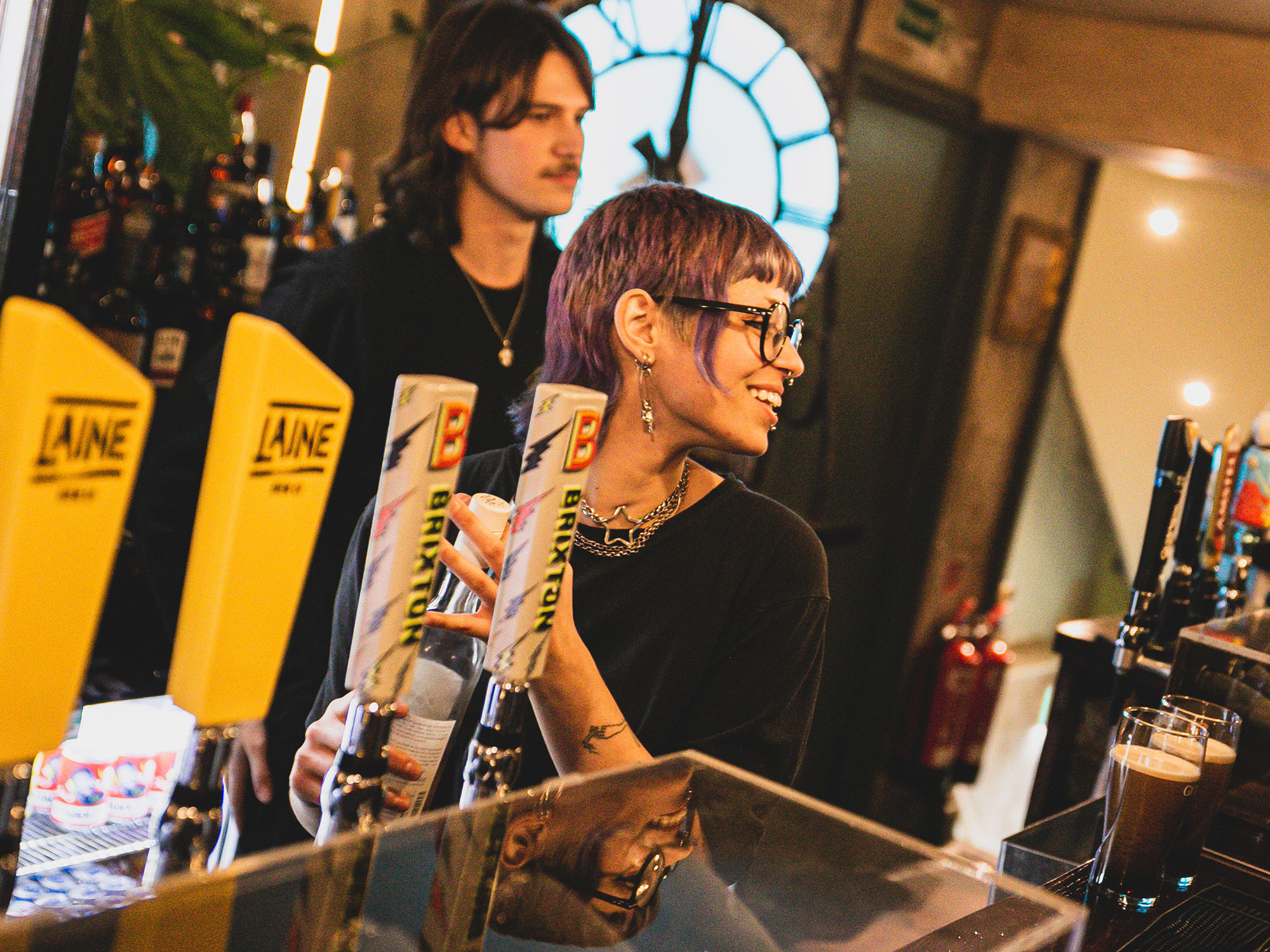 Oculist Brighton bar staff pouring a pint behind the bar with two pints of Guinness on the counter, ambient lighting and a giant lit up clock in the background.