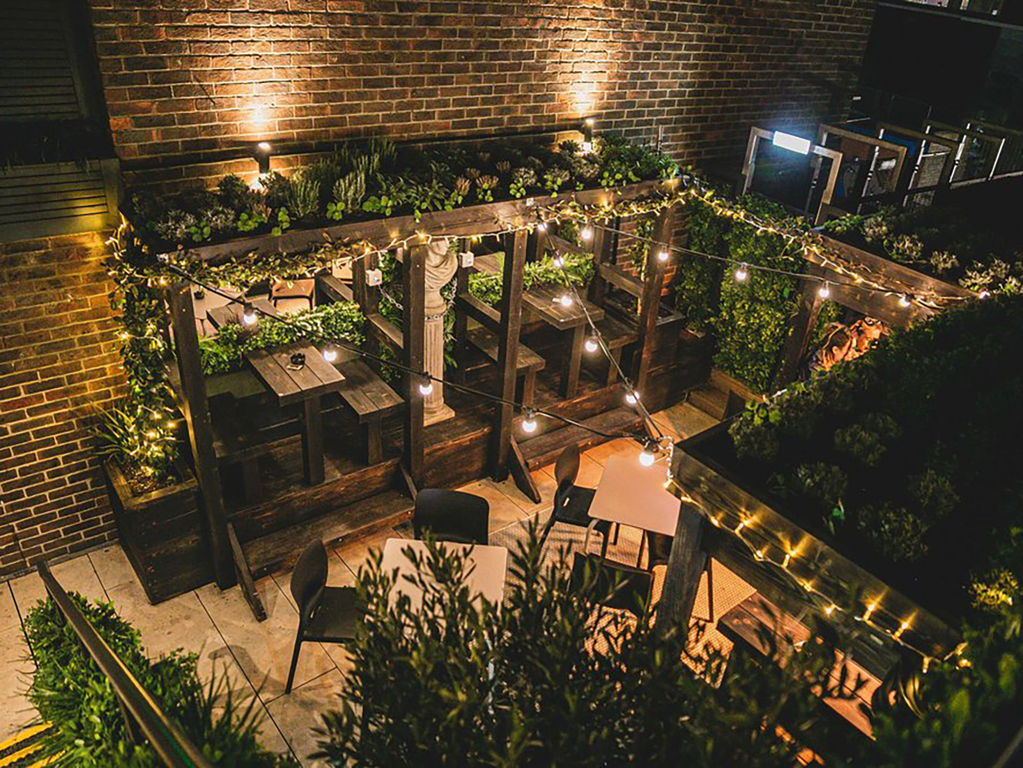 Oculist Brighton roof garden with plants and fairy lights. Night time setting, the outdoor terrace is next to a tall red brick wall. There are wooden tables under pagodas and plastic tables and chairs on the medium sized terrace. 