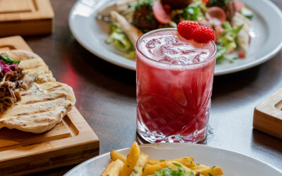 A bright red cocktail in a short glass with plates and wooden boards of food around the drink.