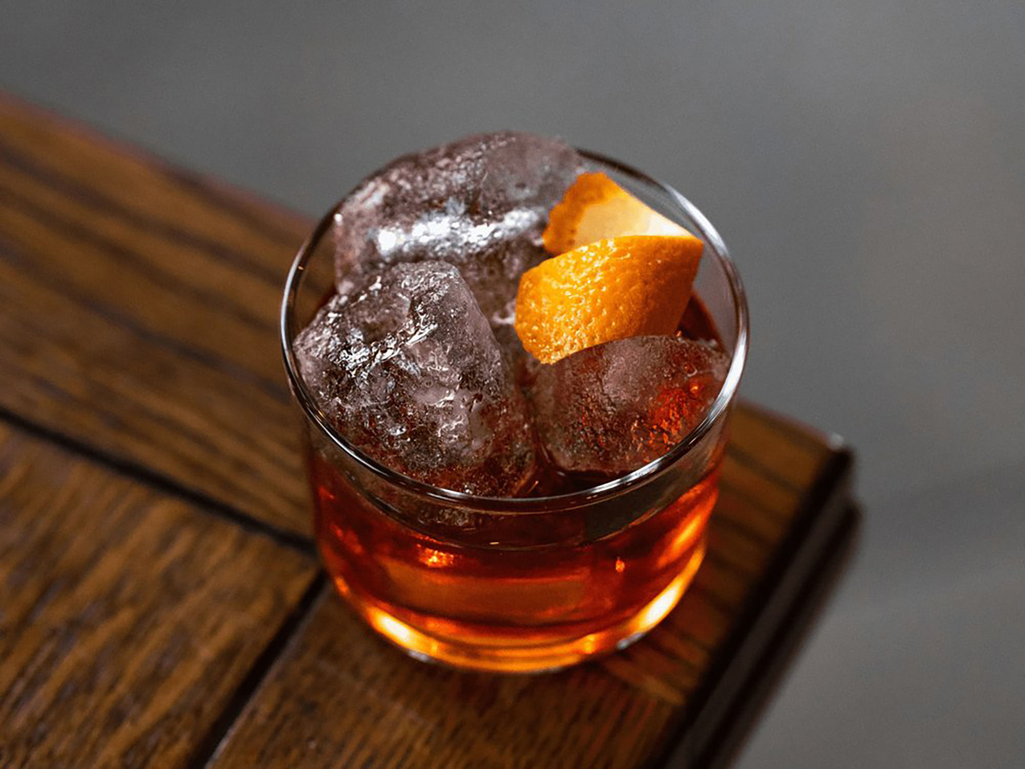 A small cocktail at Oculist Brighton. A glass of orange liquid with ice cubes, garnished with citrus peel and photographed on the corner of a wooden table.