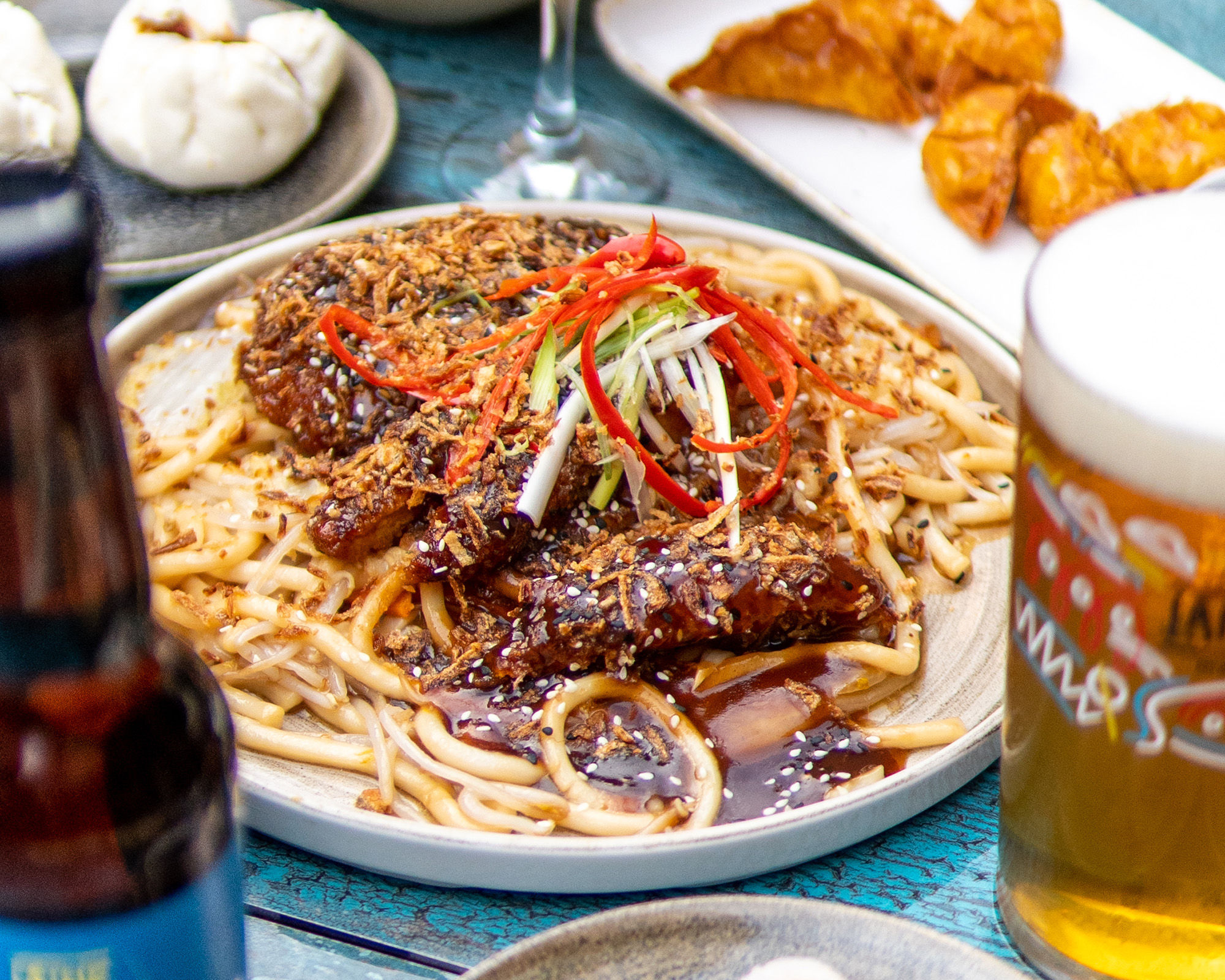 A close up shot of plate of udon noodles with a bottle and glass of beer at Oculist Brighton. The noodles have a dark sauce and are garnished with sliced red chilli and spring onions.
