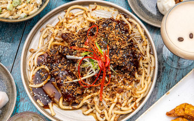 A large plate of udon noodles at Oculist Brighton with sliced red chilli and spring onions. An overhead photo, shot with small plates of dim sum surrounding the noodles on a rustic wooden blue table. Pan Asian cuisine