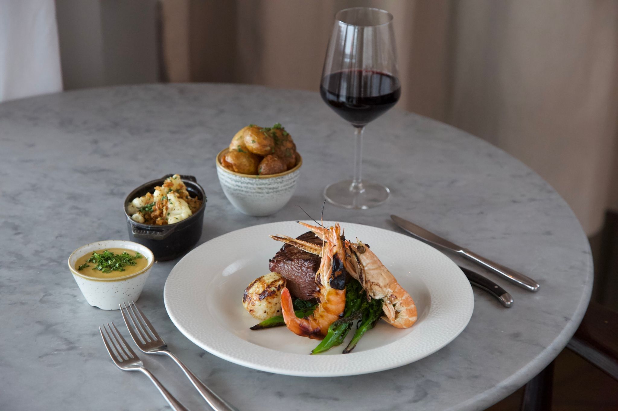 Chateaubriand, Scallop, Grilled King Prawn and Langoustine, Burnt Butter Bearnaise, Truffle Cauliflower Cheese and Crispy Garlic Potatoes served on grey table and with glass of red wine, all part of the Surf N' Turf offer