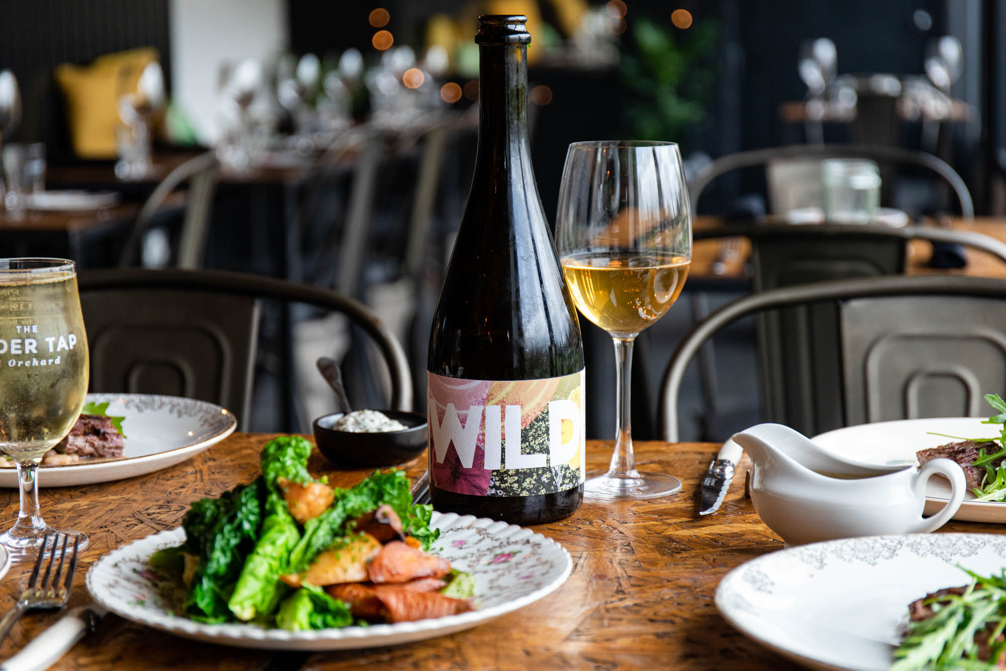A dark wooden table with a plate of food with seasonal vegetables and a glass cider with the bottle. Inside The Cider Tap Sussex with the rest of the venue out of focus in the background.