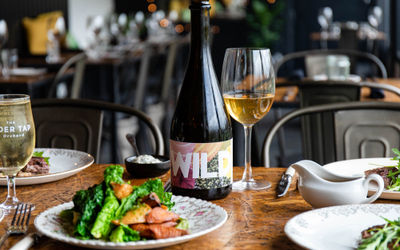 A dark wooden table with a plate of food with seasonal vegetables and a glass cider with the bottle. Inside The Cider Tap Sussex with the rest of the venue out of focus in the background.