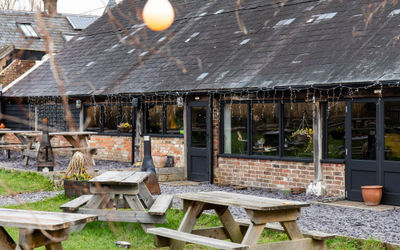 Exterior shot of The Cider Tap Sussex. A lawn with wooden tables infront of the red brick venue with a black slate roof. There are atmospheric bulb fairy lights in the garden.