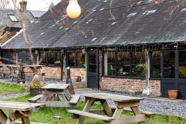 Exterior shot of The Cider Tap Sussex. A lawn with wooden tables infront of the red brick venue with a black slate roof. There are atmospheric bulb fairy lights in the garden.