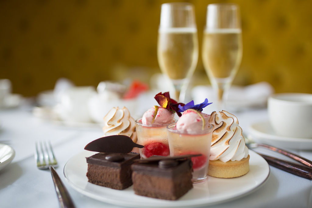 Afternoon tea at The Grand Hotel in Brighton. A tea plate with a selection of patisseries decorated with edible flowers. Two champagne flutes in the background against a yellow cushioned chair.