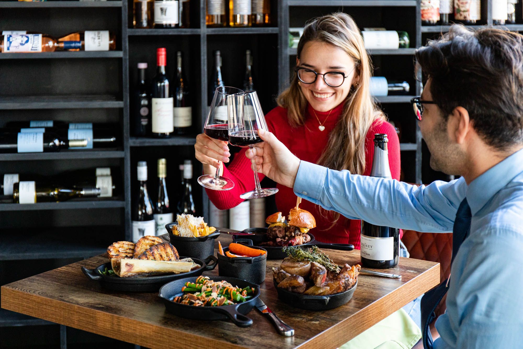 couple having a toast with glasses of red wine above their brown table laid out with various meat dishes at Carne Hove