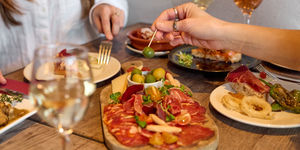 Antipasti at Tinto Taperia Spanish restaurant in Brighton - with a selection of meats on a chopping board. There's a bowl of mixed olives with two women eating food. Two glasses of white wine are on the table. Tapas Brighton.