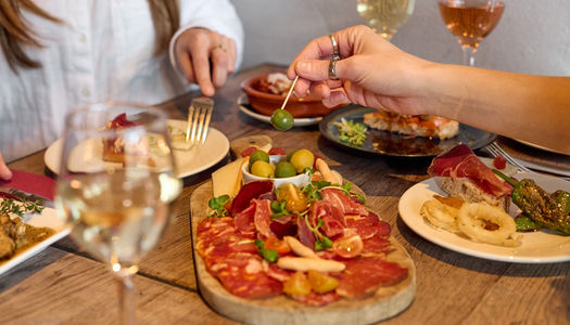 Antipasti at Tinto Taperia Spanish restaurant in Brighton - with a selection of meats on a chopping board. There's a bowl of mixed olives with two women eating food. Two glasses of white wine are on the table. Tapas Brighton.