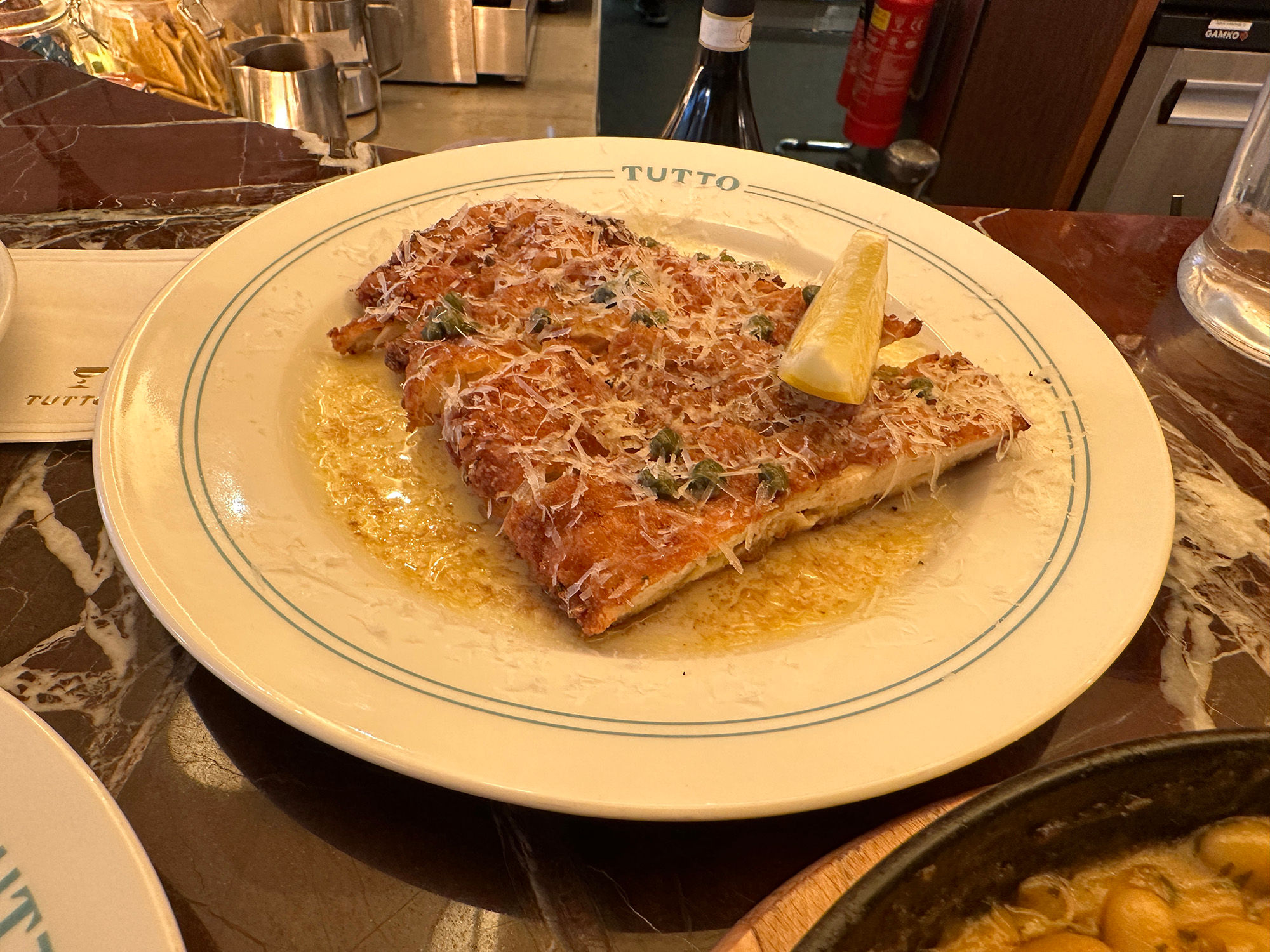 A piece of Chicken Milanese with grated parmesan and a wedge of lemon served on a Tutto branded white plate.