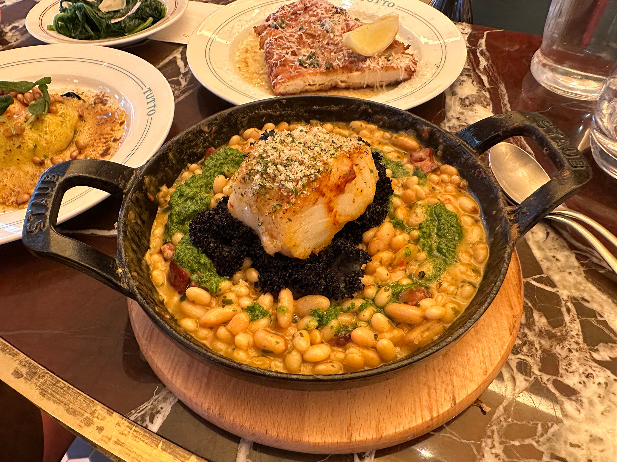 A piece of cod served with white beans in a cast iron dish on a wooden place mat. Served alongside a few Italian sharing plates.