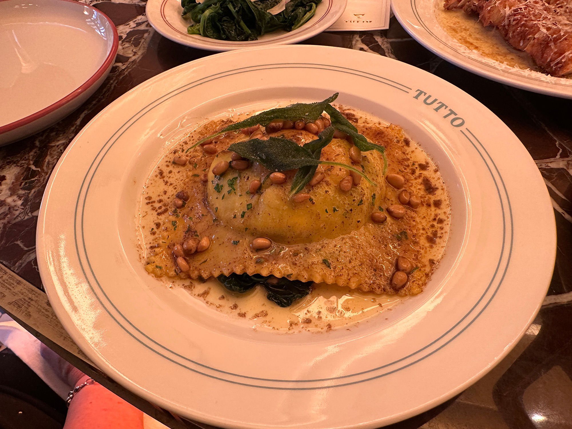 A large raviolo with crinkle edges filling a small plate. Served with toasted pine nuts and a fresh herb.