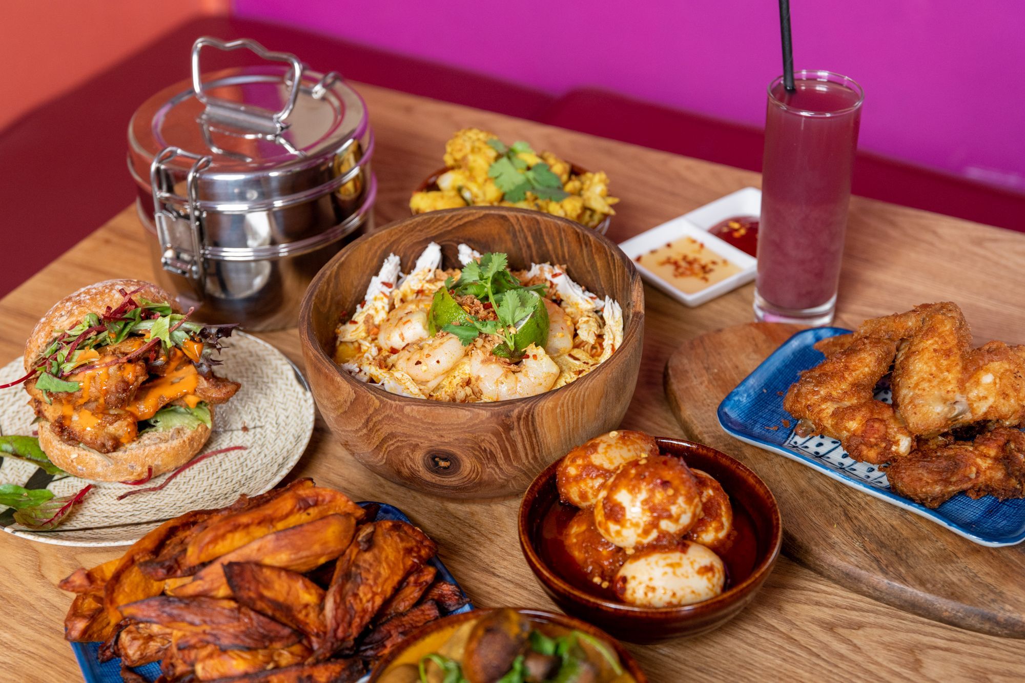 wooden brown table laid out with dishes including plantain, fried chicken, burger, fries, dumplings