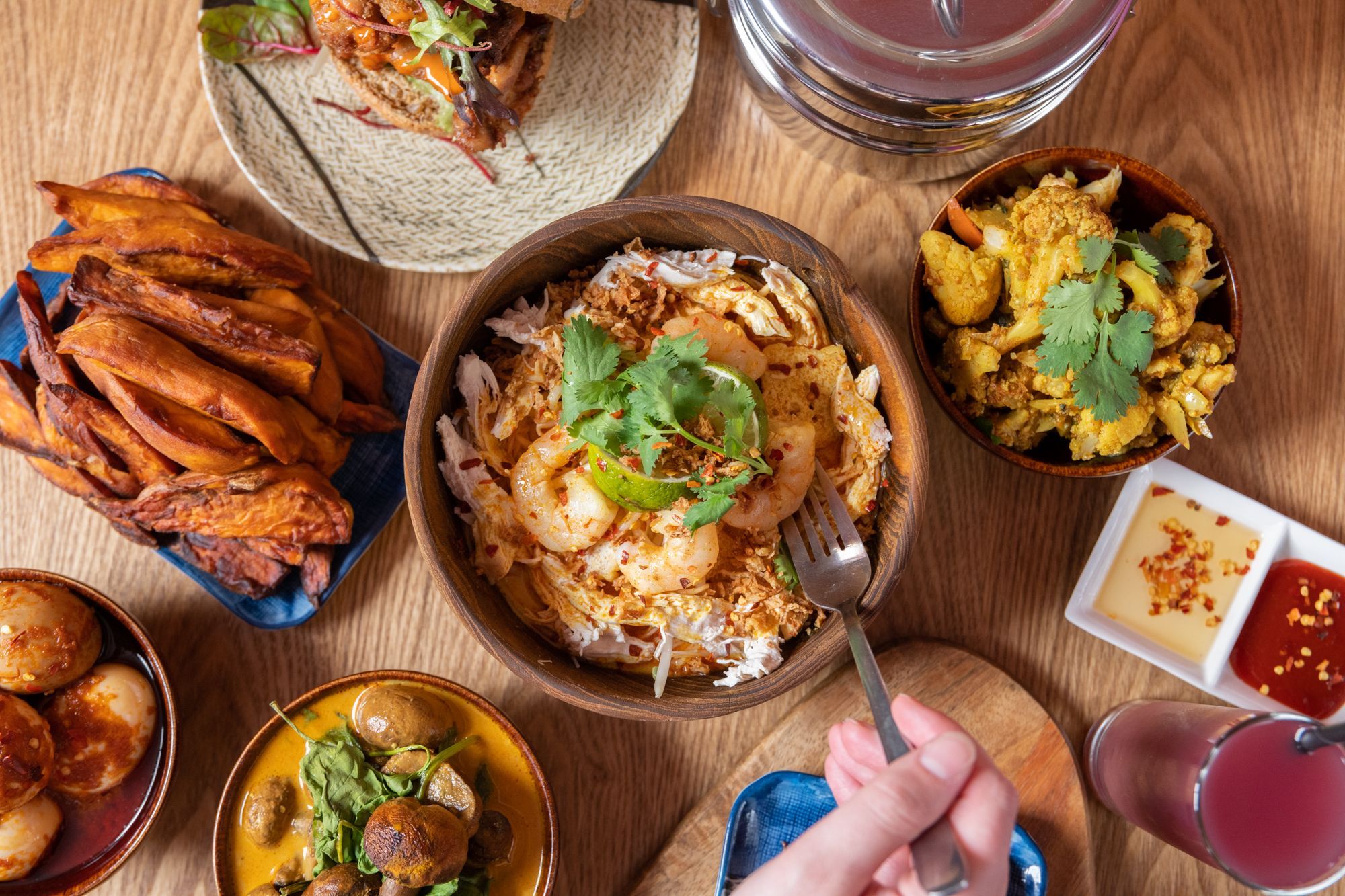 over head shot of the wooden brown table laid out with dishes including plantain, fried chicken, burger, fries, dumplings