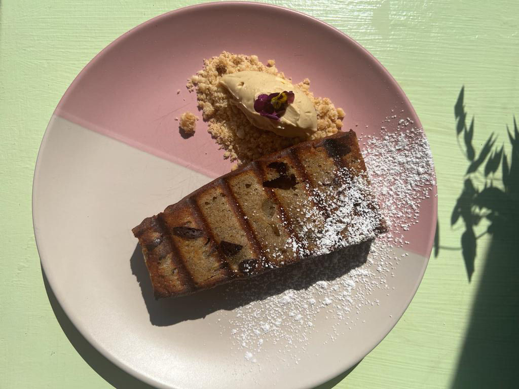 A slice of banana toast on a white and pink ceramic tea plate photographed on a green wooden table for the full Oeuf experience.