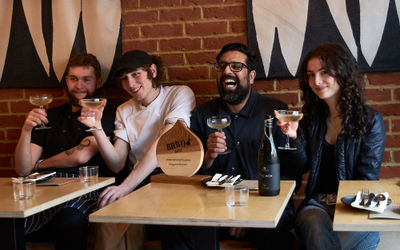 Inside Kitgum Kitchen with the team celebrating their BRAVO award with prosecco in front of a red brick wall.