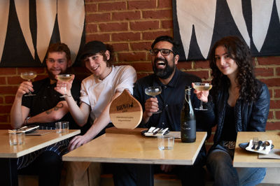 Inside Kitgum Kitchen with the team celebrating their BRAVO award with prosecco in front of a red brick wall.