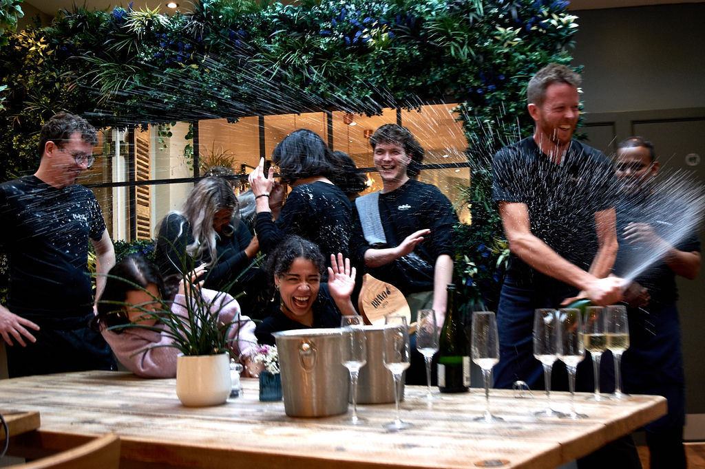 The staff at Moksha Caffe celebrating their award by spraying champagne everywhere. Champagne flutes are on the wooden table with wine coolers. Brighton brunch award. Things to do in Brighton.. January