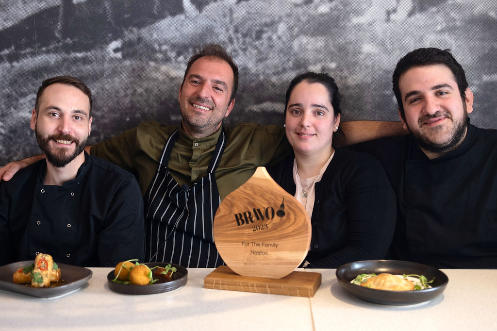 A team photo with the staff from Nostos sat at a table with plates of food and the wooden BRAVO trophy.
