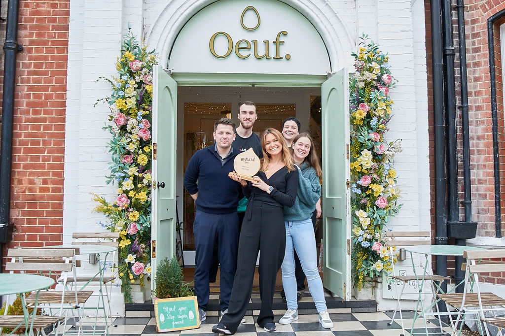 The staff at Oeuf cafe holding their wooden BRAVO trophy in front of the entrance which is decorated with flower garlands. The large Oeuf sign is above the door. Brighton brunch award