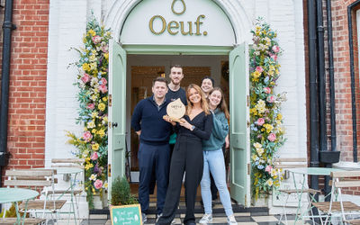 The staff at Oeuf cafe holding their wooden BRAVO trophy in front of the entrance which is decorated with flower garlands. The large Oeuf sign is above the door. Brighton brunch award