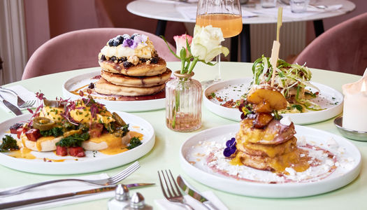 A selection of brunch dishes served on a wooden table painted light green with a small glass vase in the middle of the table with roses.