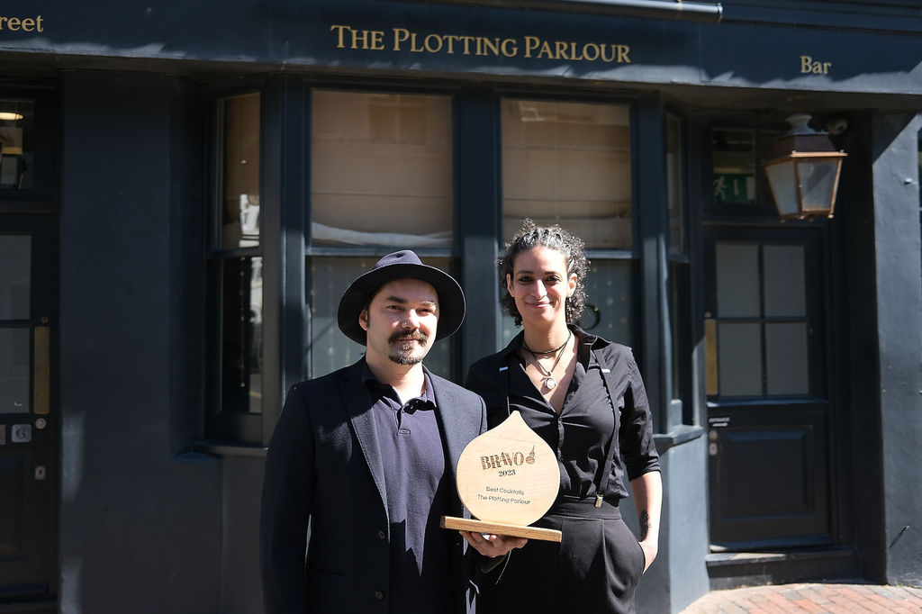 Two people holding a wooden trophy in front of The Plotting Parlour cocktail bar on a sunny day. Alcohol free Brighton