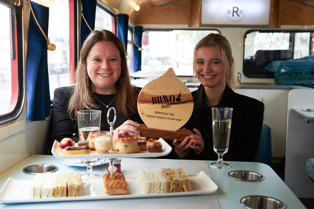 Two people holding their trophy inside a double decker bus with a three tier afternoon tea and two glasses of prosecco. Cake shop Brighton