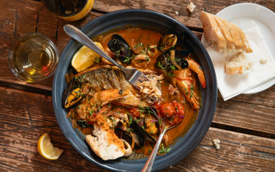 a black bowl of fish stew sitting on a wooden slatted table. side dish with bread. a glass of white wine.