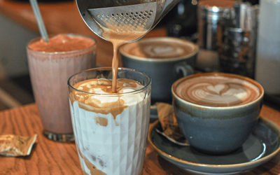An iced coffee being made a Vios Cafe which is being served in a glass tumbler with cups of coffee in the background which have latte art hearts on them.