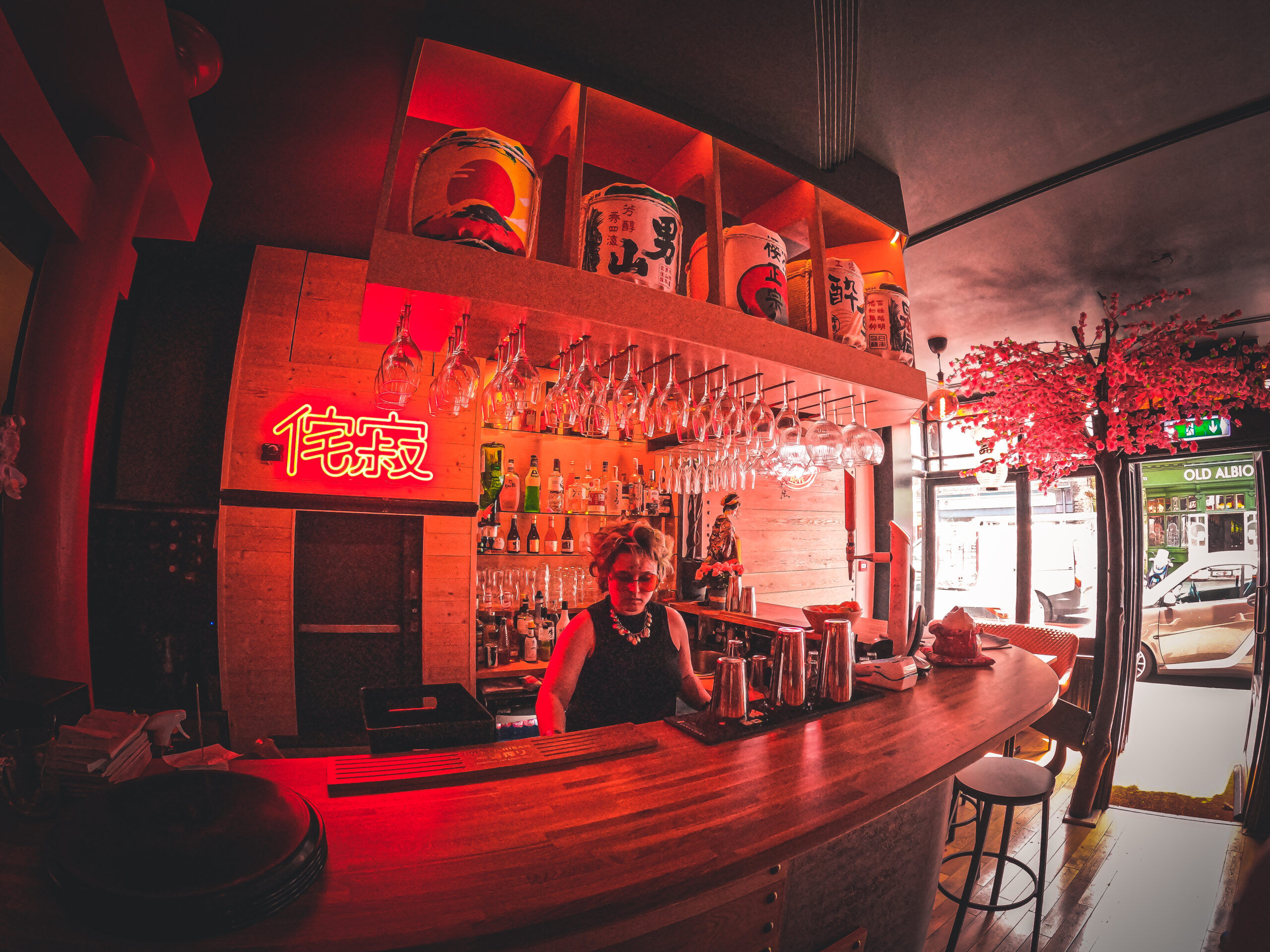 The bar area at Wabi Sabi with a bartender wearing sunglasses. A neon light red light lights up the area.