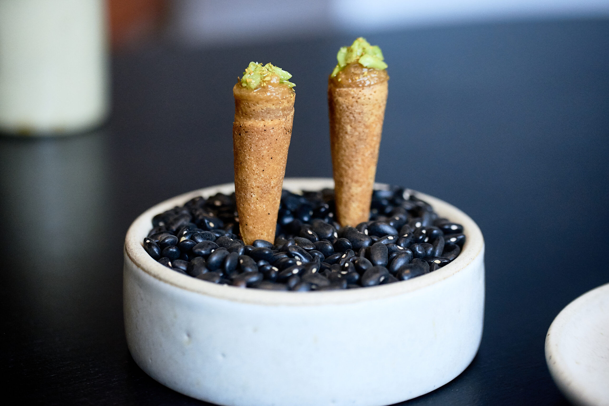 Crispy pancake cones with a cheese mousse and fennel jam displayed on dried black beans in a white ceramic dish.