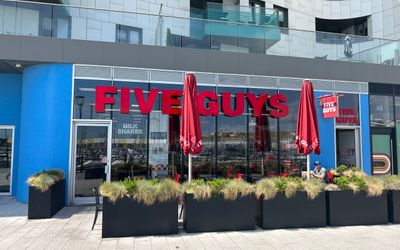 The facade of Five Guys Pizza on Brighton Marina harbour area. A blue facade with red signage and red umbrellas next to green hedges which frame the alfresco area.
