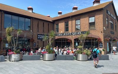 the red building of The West Quay on Brighton Marina. Alfresco drinkers and diners, with trees dotted around in big pots. A blue sky in the background