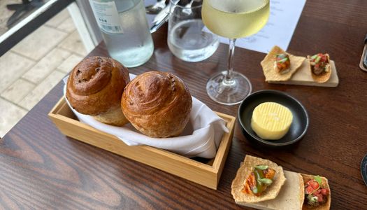 two bread rolls served on the wooden plate together with butter in the small black bowl and glass of white at the fig tree