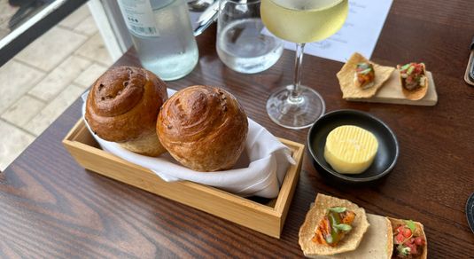 two bread rolls served on the wooden plate together with butter in the small black bowl and glass of white at the fig tree